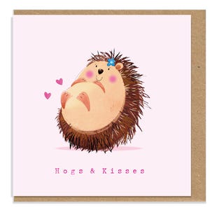 May include: A square greeting card with a light pink background featuring a cartoon hedgehog with a blue flower on its head, pink cheeks, and two small pink hearts. The text "Hogs & Kisses" is printed in pink at the bottom of the card.