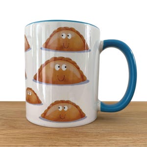May include: A white ceramic mug featuring a repeating pattern of brown, crescent-shaped empanada illustrations with simple cartoon faces, including two eyes and a smile. The mug has a bright blue handle and a matching blue interior rim.