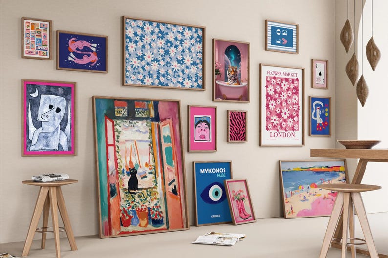 Gallery wall featuring a curated collection of diverse prints and artwork.  The pieces include floral patterns in pink and blue, a queen of hearts playing card, a cat in a window scene, an evil eye, and various abstract designs.  The color palette is vibrant, with pinks, blues, and greens dominating. The prints are framed in light wood and arranged in a visually appealing, eclectic style.  The overall theme is modern and playful.