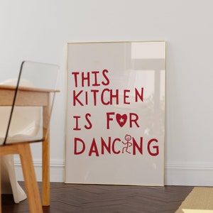 Puede incluir: Un póster enmarcado blanco con texto rojo que dice "This kitchen is for dancing" con un corazón rojo y una figura de palo bailando.