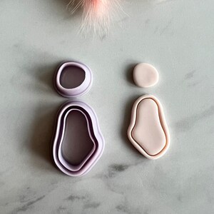 Puede incluir: Conjunto de cuatro cortadores de galletas de plástico morado y rosa con forma de formas orgánicas abstractas. Los cortadores están dispuestos sobre una superficie de mármol blanco.