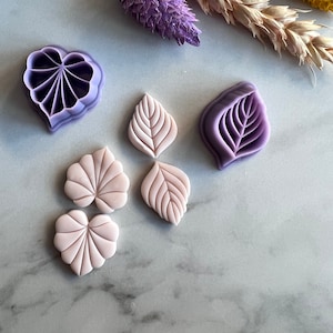 Puede incluir: Un conjunto de cinco cortadores de hojas y flores de arcilla polimérica de color morado y rosa. Los cortadores están dispuestos sobre una superficie de mármol blanco.