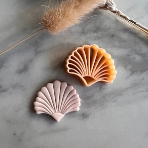 Puede incluir: Dos moldes de silicona en forma de concha de mar de color rosa y naranja sobre una superficie de mármol. Los moldes son para hornear o manualidades.