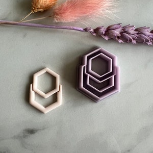 Puede incluir: Dos juegos de cortadores de galletas geométricas, un juego es blanco y el otro es morado. Ambos juegos tienen forma de hexágonos con una doble capa.