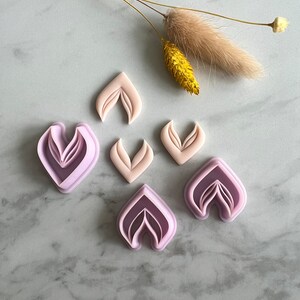 Op de afbeelding: Zes paarse plastic bloemvormige koekjesuitstekers op een wit marmeren oppervlak. De uitstekers hebben de vorm van een bloem met meerdere lagen bloemblaadjes.