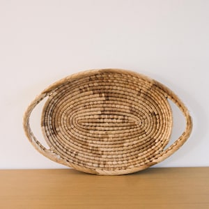 May include: An oval-shaped woven basket featuring a light tan, natural fibre construction. The basket has a coiled base and a thick, braided rim that extends outwards on both sides to form integrated handles, creating a symmetrical design.