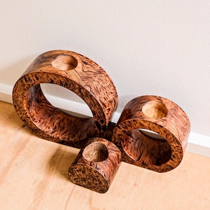 May include: Three wooden candle holders in different sizes. They are made from dark, patterned wood with a circular shape and a central hole for a candle. The wood grain is visible. The candle holders are arranged on a light-coloured surface.