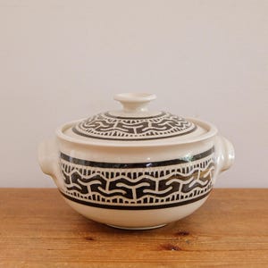 May include: A white ceramic lidded serving bowl with a black geometric pattern. The bowl has two handles and a knob on the lid. The design is a repeating pattern of black shapes on a white background. The bowl is sitting on a wooden surface.
