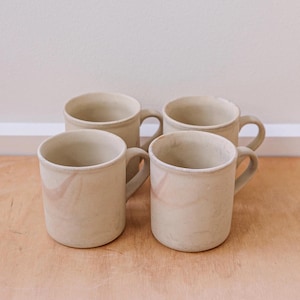 May include: Four beige ceramic mugs with handles are displayed on a wooden surface. Each mug has a cylindrical shape with a slightly flared rim and a curved handle. Subtle pinkish-brown swirls decorate the exterior of the mugs, adding visual interest.