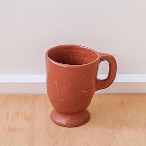 May include: A terracotta-coloured mug with a rounded body and a small handle. The mug has a pedestal base and a slightly textured surface, suggesting a rustic, artisanal quality. The mug is designed for hot beverages.