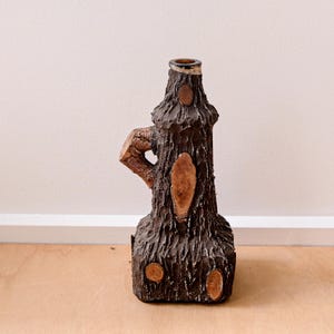 May include: A decorative bottle shaped like a tree trunk. The brown bottle has a textured surface with wooden accents and a handle. The top of the bottle is a darker brown colour.