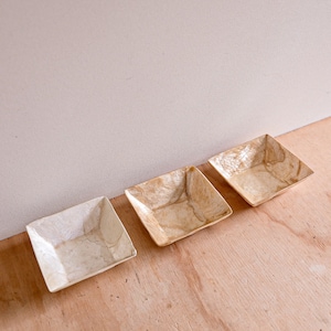 May include: Three square-shaped decorative bowls made of natural materials. The bowls are light brown with a white marbled pattern. The bowls are arranged in a row on a wooden surface.