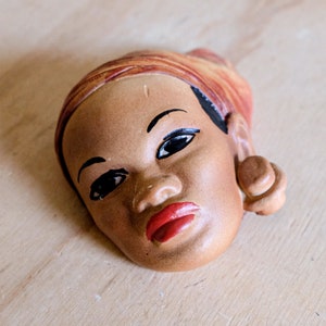 May include: A ceramic wall sculpture of a woman's face with a red and orange head wrap and a large brown earring. The woman has brown skin and black eyes.