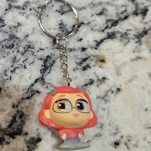May include: A keychain featuring a cartoon character with red hair, glasses, and a red shirt. The figure is attached to a silver chain and ring. The background is a speckled gray and white surface.