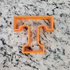 May include: An orange plastic cookie cutter in the shape of the letter "T". The cutter is a single piece with a rectangular top and two curved vertical lines extending downwards, connected at the bottom.