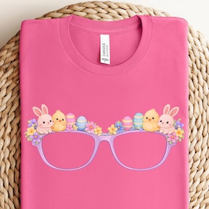 Puede incluir: Una camiseta rosa con un gráfico de gafas moradas decoradas con elementos de Pascua. El diseño incluye conejitos, pollitos, flores y huevos de Pascua. La camiseta se muestra sobre una superficie circular tejida.