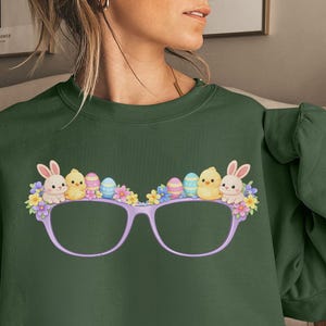 Puede incluir: Una sudadera verde militar con un diseño lúdico de Pascua. El diseño presenta gafas decoradas con conejitos, pollitos, huevos coloridos y flores. El aspecto general es festivo y caprichoso, perfecto para la temporada de primavera.
