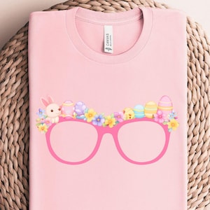 May include: Pink t-shirt featuring a graphic of pink glasses with an Easter-themed design. The design includes a bunny, Easter eggs, chicks, and flowers. The shirt is displayed on a woven, circular surface.