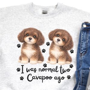 May include: A light gray sweatshirt featuring two brown and white Cavapoo puppies with paw prints. The text "I was normal two Cavapoo ago" is printed below the puppies. The sweatshirt is paired with blue jeans, white shoes, and a gray travel mug.