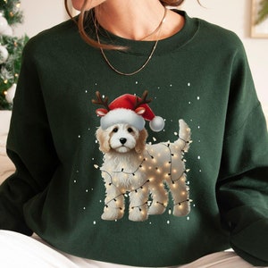 May include: A forest green sweatshirt with a white dog wearing a red Santa hat and surrounded by white Christmas lights. The dog is wearing a red Santa hat and has a white beard.