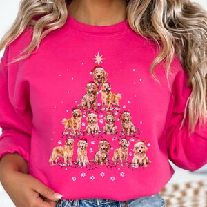 Può includere: Una felpa rosa acceso con un design a forma di albero di Natale composto da cuccioli di golden retriever con cappelli da Babbo Natale e luci a catena. Una stella in cima all'albero. La felpa è a girocollo.