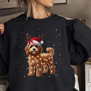 May include: A black sweatshirt with a Christmas-themed design featuring a brown dog wearing a Santa hat and reindeer antlers, surrounded by fairy lights. The dog is standing on a snowy background.