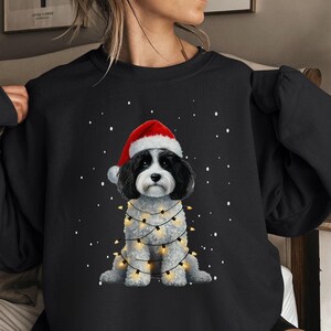 May include: Black sweatshirt featuring a white and black dog wearing a red Santa hat and surrounded by Christmas lights. The dog is sitting against a snowy backdrop.