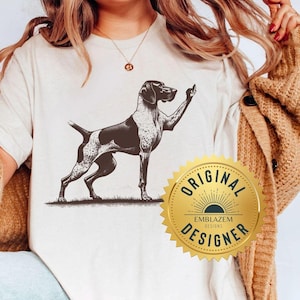 May include: A white t-shirt with a black and white illustration of a German Shorthaired Pointer dog giving the middle finger. The shirt has a gold seal with the text "Original Emblazem Designs Designer" on it.