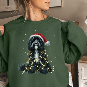 May include: A dark green sweatshirt featuring a black and white dog wearing a Santa hat and surrounded by Christmas lights. The dog is sitting on a white background with snowflakes.