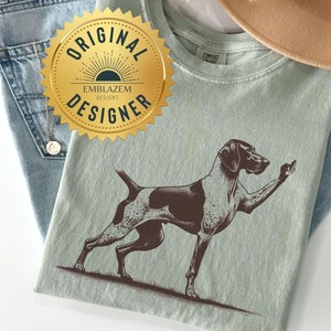 May include: A light green t-shirt with a brown and white illustration of a dog giving the middle finger. The shirt has a gold label that says "Original Designer" with a sunburst design.