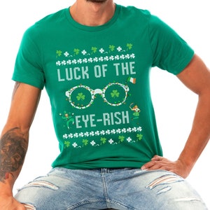 May include: Green t-shirt with the text "LUCK OF THE EYE-RISH" in white. The design includes glasses with shamrocks, a leprechaun, and an Irish flag. The shirt is a short-sleeved crew neck.