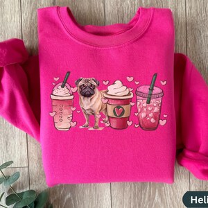 May include: Pink sweatshirt with a graphic of a pug dog standing in front of three coffee cups with hearts. The cups are filled with coffee, whipped cream, and iced coffee.