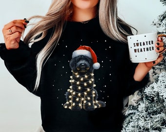 May include: Black sweatshirt featuring a black dog wearing a Santa hat and Christmas lights. The text "Sweater Weather" is on a white mug in the image.