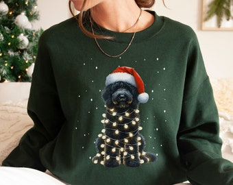 Könnte beinhalten: Ein dunkelgrüner Pullover mit einem schwarzen Hund, der einen Weihnachtsmann-Hut trägt und von Weihnachtslichtern umgeben ist. Der Hund sitzt auf einem weißen Hintergrund mit Schneeflocken.