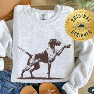 Pode incluir: Uma camisa de moletom cinza com uma ilustração em preto e branco de um cão Braco Alemão de Pelo Curto apontando. A camisa de moletom tem gola redonda e mangas compridas. O texto "ORIGINAL DESIGNER" está em um adesivo circular dourado na camisa de moletom.