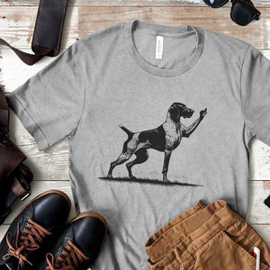 Pode incluir: Uma camiseta cinza com uma ilustração em preto e branco de um Braco Alemão de Pelo Curto mostrando o dedo do meio. O texto "Athletic Heather" está impresso no canto inferior direito da camiseta.