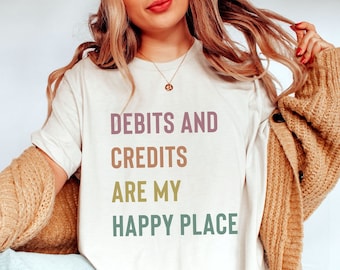 Puede incluir: Camiseta blanca con el texto "Debits and Credits are my Happy Place" en letras de colores.
