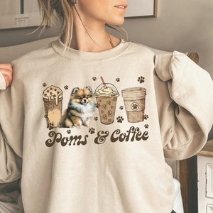 Puede incluir: Una sudadera beige con un gr&aacute;fico marr&oacute;n de un perro Pomerania y tres tazas de caf&eacute;. El texto "Poms & Coffee" est&aacute; impreso debajo del gr&aacute;fico.