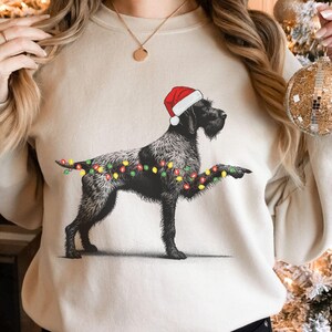 May include: Sand-coloured sweatshirt with a black dog illustration wearing a Santa hat and Christmas lights. The dog is detailed, with lights wrapped around its body and paw. A festive and cosy design for the holiday season.