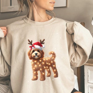 May include: A beige sweatshirt featuring a Christmas-themed illustration of a brown dog wearing a Santa hat and reindeer antlers. The dog is surrounded by white lights.