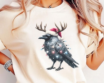 May include: A cream colored t-shirt with a graphic of a black crow wearing a Santa hat and reindeer antlers with Christmas lights wrapped around it. The crow is standing on a snowy background.