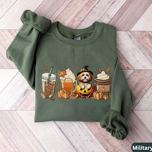 May include: A military green sweatshirt with a cute dog wearing a witch hat and holding a pumpkin. The dog is surrounded by fall-themed drinks and pumpkins.