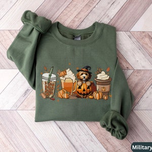 May include: A military green sweatshirt with a graphic of a dog wearing a witch hat and holding a pumpkin. The graphic also includes fall-themed elements like coffee, pumpkin spice, and leaves.