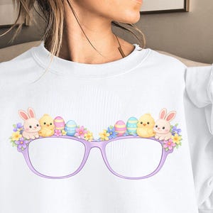 Puede incluir: Sudadera blanca con un diseño temático de Pascua. El gráfico incluye unas gafas moradas adornadas con conejitos de dibujos animados, pollitos, flores y huevos de Pascua. La palabra "White" es visible en la esquina inferior derecha.