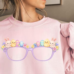 Puede incluir: Una sudadera rosa claro con un diseño caprichoso de gafas moradas adornadas con elementos temáticos de Pascua. Las gafas están decoradas con pollitos de dibujos animados, conejitos, flores y coloridos huevos de Pascua. La sudadera es de color rosa claro liso.