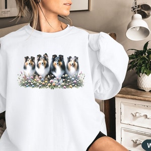 Puede incluir: Sudadera blanca con un gr&aacute;fico de cinco Shetland Sheepdogs sentados en fila en un campo de flores. Los perros son todos negros y blancos con pelaje esponjoso.