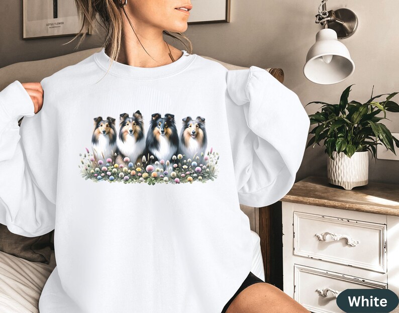 Puede incluir: Sudadera blanca con un gr&aacute;fico de cinco Shetland Sheepdogs sentados en fila en un campo de flores. Los perros son todos negros y blancos con pelaje esponjoso.