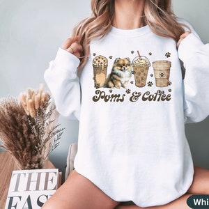 Puede incluir: Sudadera blanca con un perro pomerania marr&oacute;n y tres tazas de caf&eacute; con el texto "Poms & Coffee".