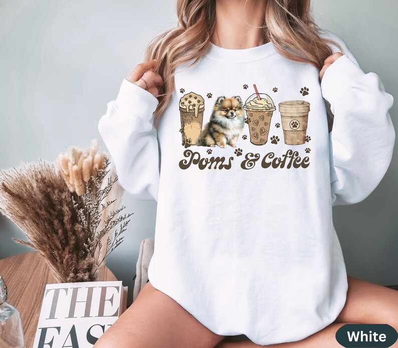 Puede incluir: Sudadera blanca con un perro pomerania marr&oacute;n y tres tazas de caf&eacute; con el texto "Poms & Coffee".