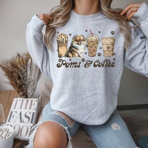 Puede incluir: Sudadera gris con un gr&aacute;fico de un perro Pomerania y tres tazas de caf&eacute;. El texto "Poms & Coffee" est&aacute; impreso en la sudadera.
